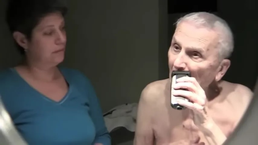 Older adult using an electric shaver in front of a mirror while another person stands nearby