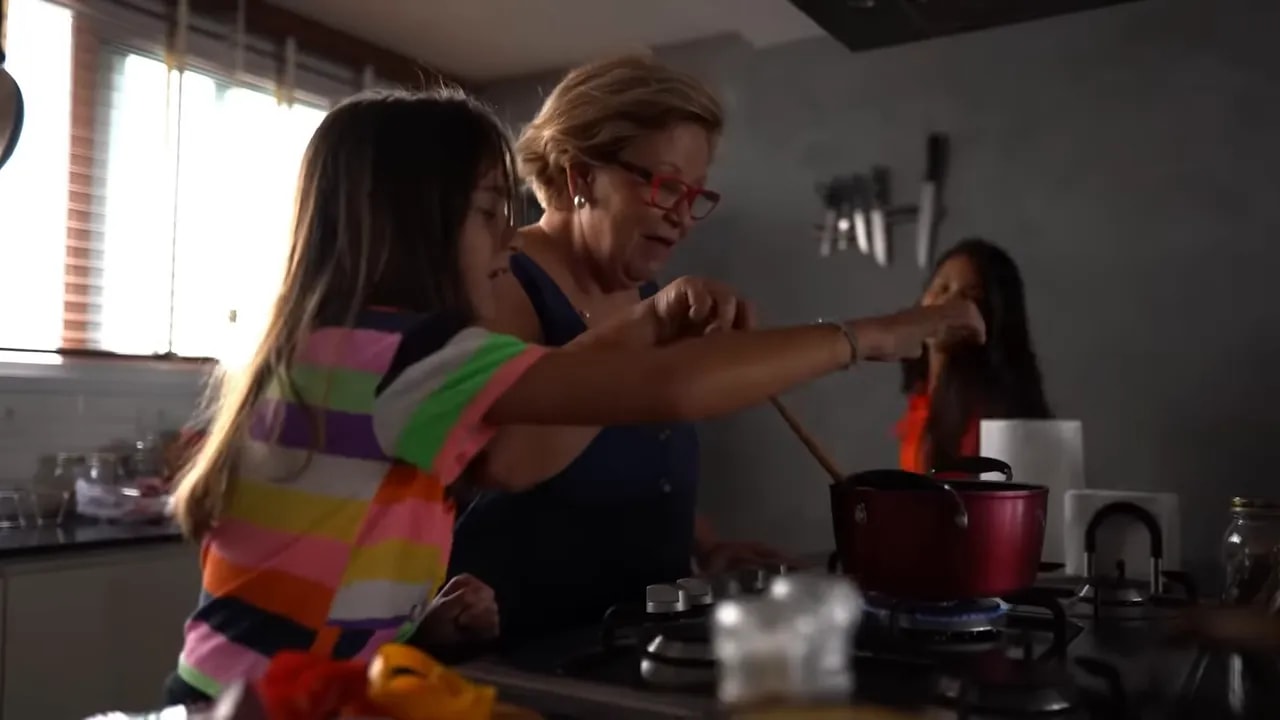 older woman and two children cooking together at a kitchen stove with a pot and utensils