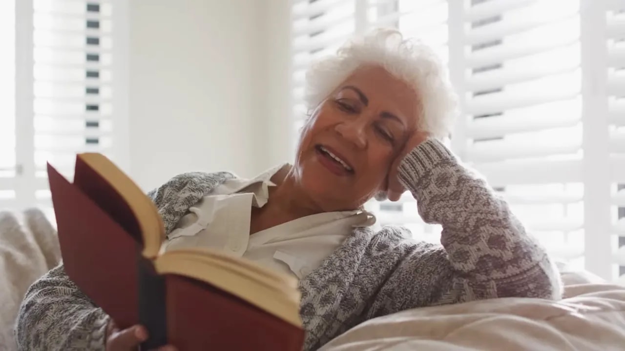 older woman reclining on a couch reading a large open book near bright window blinds