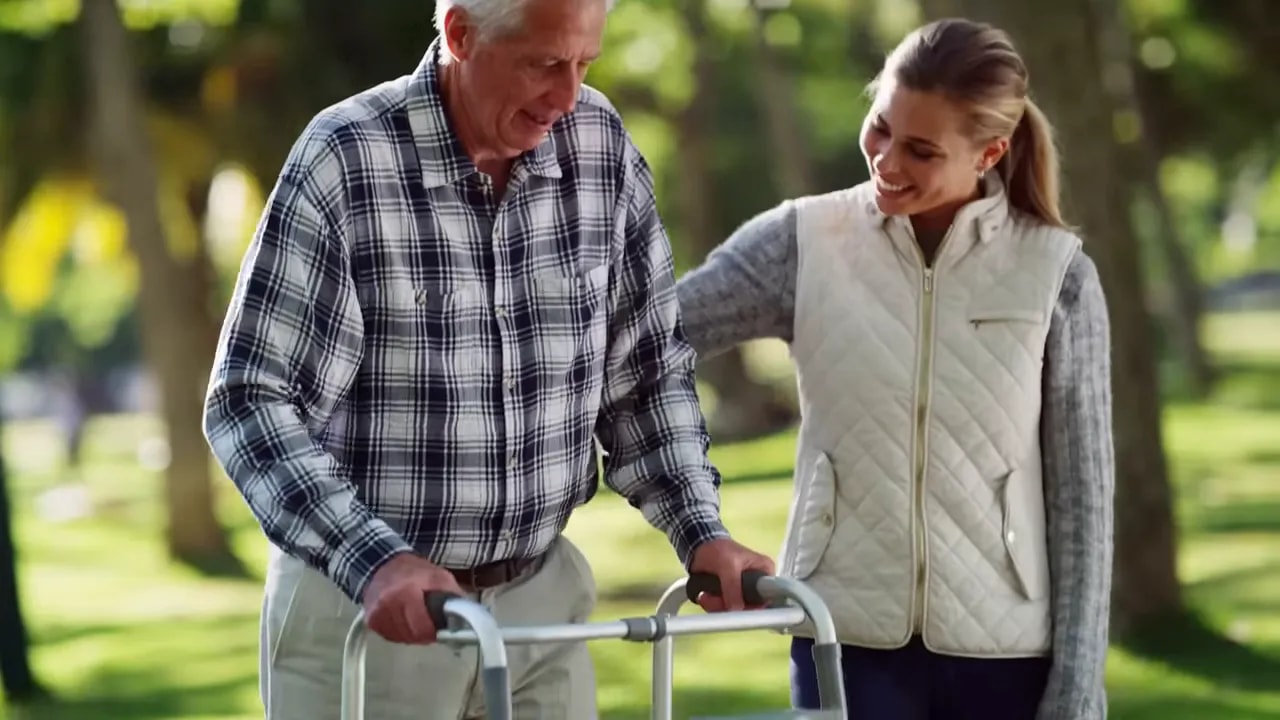older man using a walker outdoors in a park while a younger woman walks beside him for support